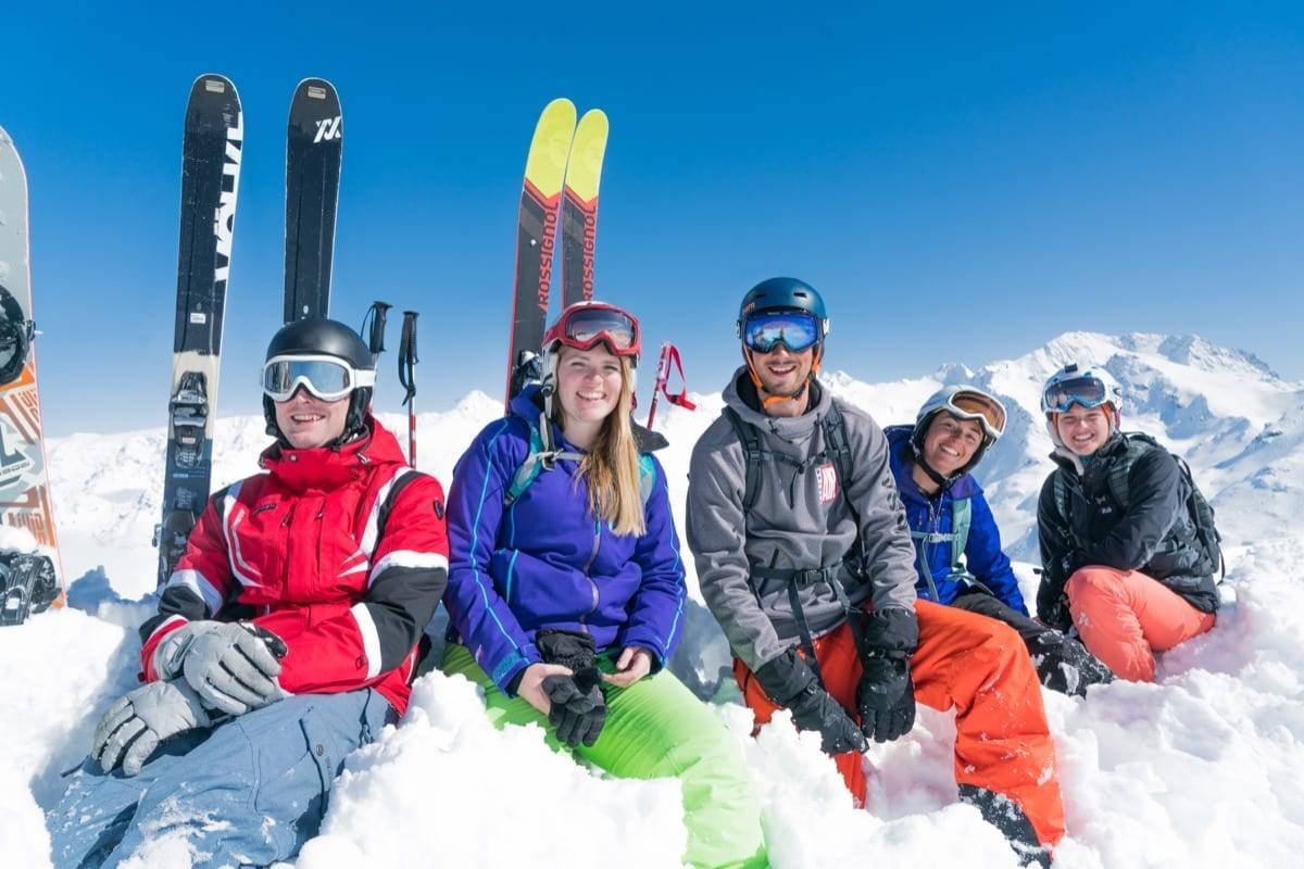 A group of friends smiling as they take a break with their skis, sitting in snow