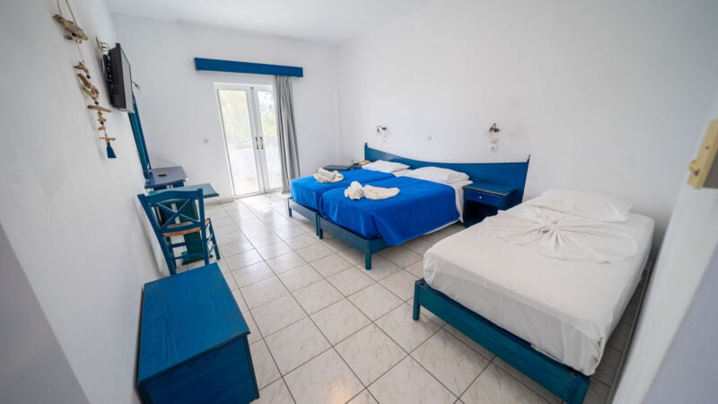 Hotel room with a mirror and flatscreen TV on the wall, two twin beds pushed together to form a double bed with a blue blanket and towels on top and a single twin bed with white bed linen.