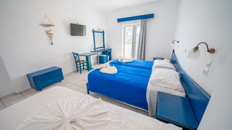 Hotel room with a mirror and flatscreen TV on the wall, two twin beds pushed together to form a double bed with a blue blanket and towels on top and a single twin bed with white bed linen.