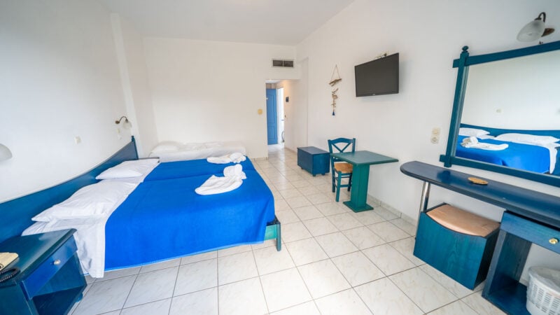 Hotel room with a mirror and flatscreen TV on the wall, two twin beds pushed together to form a double bed with a blue blanket and towels on top and a single twin bed with white bed linen.