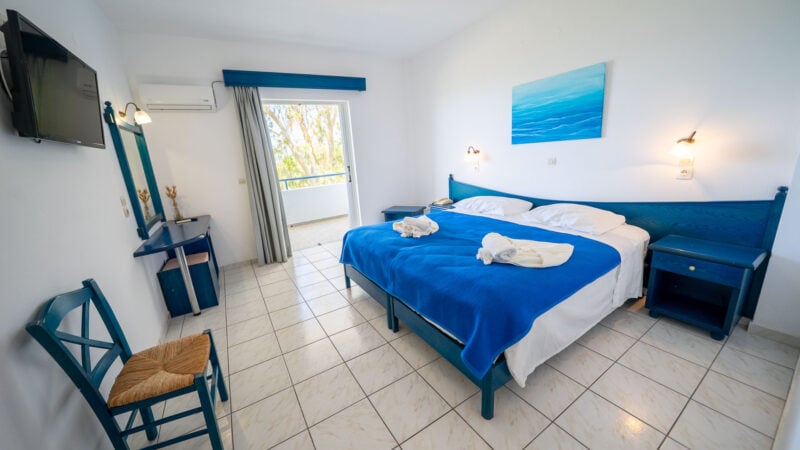 An Upper-Level Room with two twin beds together to form a double bed with a blue blanket and towels on top in a white tiled hotel room with a door leading to the balcony.