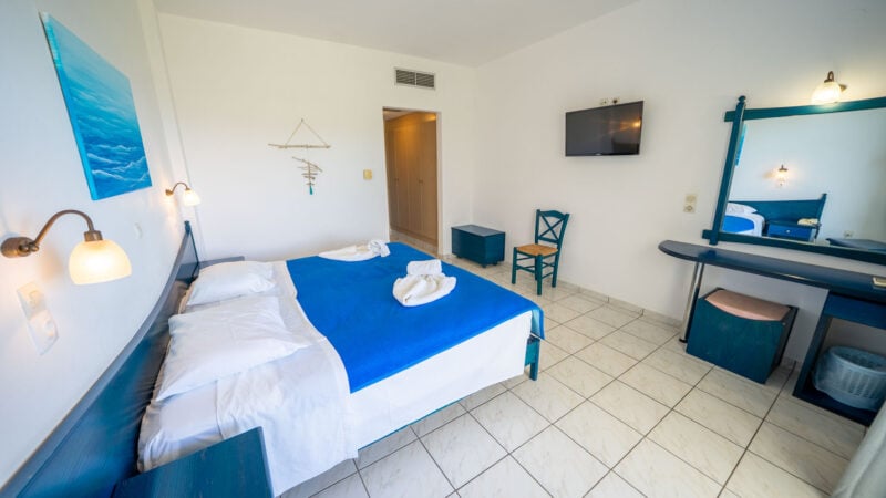 An Upper-Level Room with two twin beds together to form a double bed with a blue blanket and towels on top in a white tiled hotel room with a door leading to the balcony.