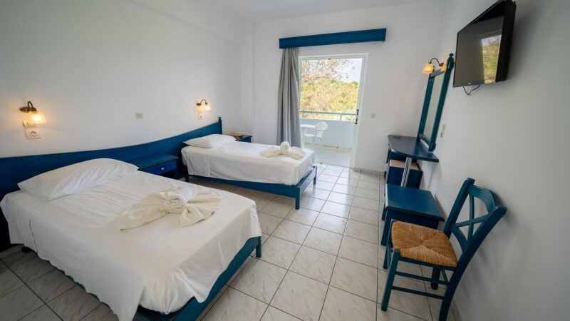 An Upper Level Room with two separated twin beds with white bed linen and towels on top of the beds. Blue chair with wicker seat and mirror on the wall.