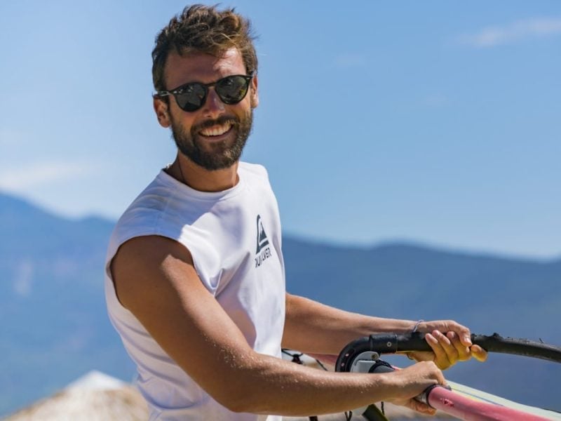 A man smiling while carrying a windsurf sail on the beach outside the Zefiros Beach Hotel in Samos, Greece